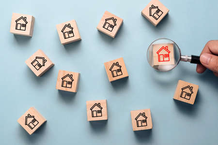 Hand holding magnifier glass to searching red house icon on wooden cube block among black house icon. Finding the best house and real estate for family concept.の写真素材