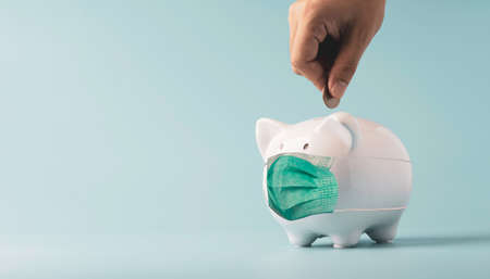 Hand putting coin to white piggy bank saving which wearing surgical mask on blue background and copy space , Money saving for future investment and retirement concept.の写真素材