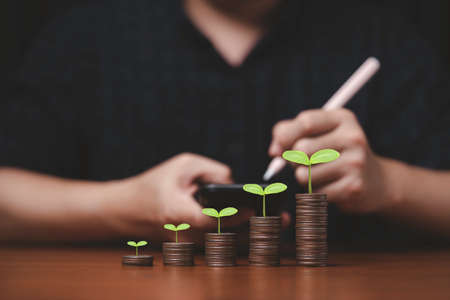 Plant growth on increasing coins stacking and businessman writing on smartphone for record and summary saving of growth profit and interest rating from investment concept.の写真素材