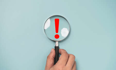Hand holding magnifier glass with red triangle caution warning sign for notification error and...