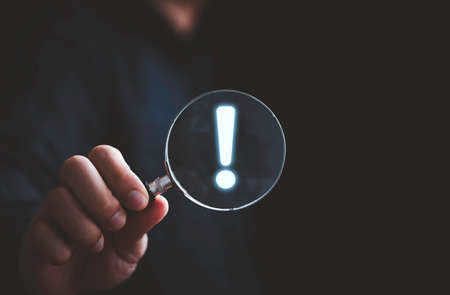Hand holding magnifier glass with red triangle caution warning sign for notification error and maintenance concept.の写真素材