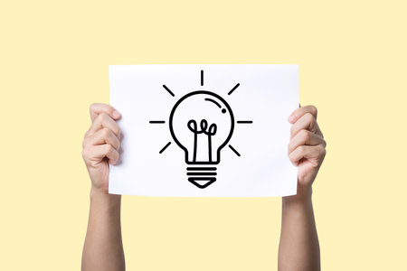 A person holding a white sheet of paper featuring a drawn light bulb, symbolizing creativity, ideas, and innovation against a soft yellow background.の写真素材