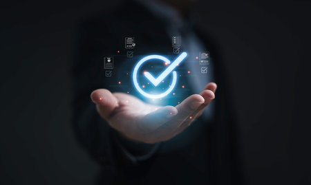 Businessman hand holding glowing check mark symbol with digital document icons representing approval process quality control verification and business success on dark backgroundの写真素材