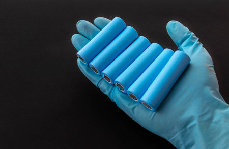 Gloved hand holds blue 18650 lithium-ion battery packs for electrical appliances and devices on black background.の写真素材
