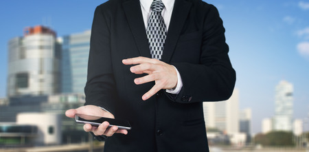 Businessman Holding Smartphone in Hand With Hand Gesture Over The Phones Screen With Business City and Corporate Buildings In Backgroundの写真素材