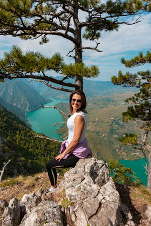 Woman hiker at mountain viewpoint resting on a rock with lake and canyon in the distance