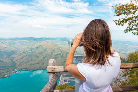 Woman hiker at mountain viewpoint looking throgh touristic telescope at the lake and canyon in the...