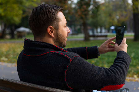 Man with smartphone in his hand taking selfie while sitting on the bench in the park at autumn