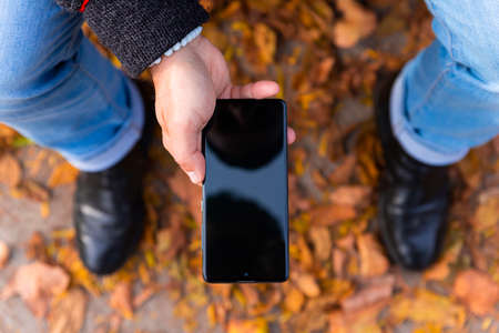 Man holding smartphone outdoors with yellow autumn leaves on the groundの写真素材