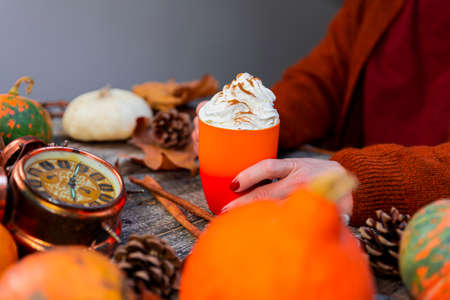 Coffee latte with whipped cream and cinnamon on top held in womans hands on a wooden background of pumpkins, autumn leaves. Fall and Thanksgiving conceptの写真素材