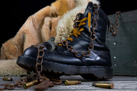 Leather boot with rusted chains and bullet shells with military gas mask and fur coat in background resting on wooden tableの写真素材