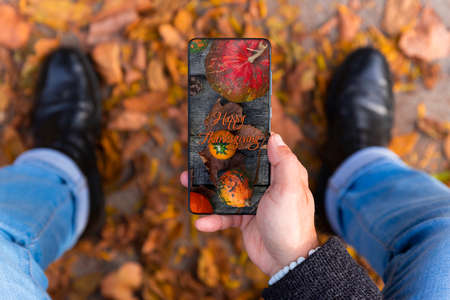 Man holding smartphone with Happy Thanksgiving photo message on the screen outdoors in a park full of autumn leaves on the groundの写真素材