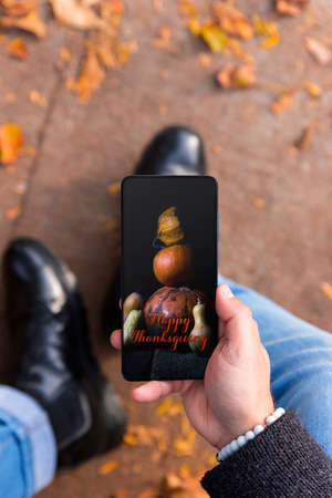 Man holding smartphone with Happy Thanksgiving photo message on the screen outdoors in a park full of autumn leaves on the groundの写真素材
