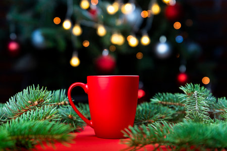 Delicious fresh festive morning coffee in a red cup on the red surface with Christmas tree lights and spruce branches in the backgroundの写真素材