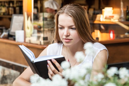 smiling girl sitting at street cafe summer terrace with flowers with morning coffee and reading bookの写真素材