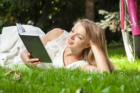 Attractive woman lays on green grass reading book in city park with pink bicycle behind herの写真素材