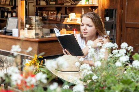 smiling girl sitting at street cafe summer terrace with flowers with morning coffee and reading book