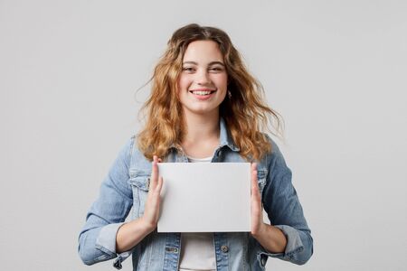 young girl with long flowing hair in a denim jacket holds a blank white card with both hands, looks...