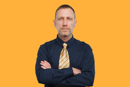 Man in a dark blue shirt and a bright yellow tie and with arms crossed on an orange backgrounds.の写真素材