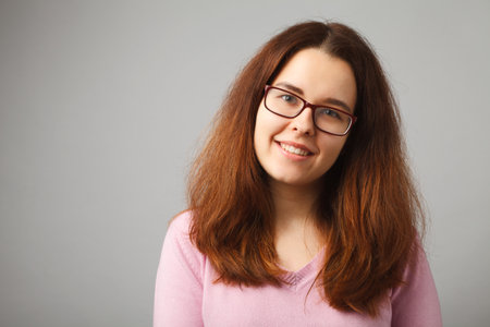 Redhead long-haired girl with glasses smiles, looking at the camera. Studio portrait of a girl on a solid light background.の写真素材