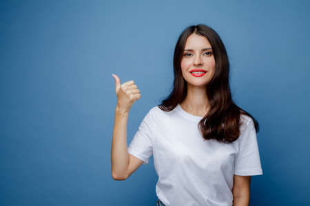 Brunette woman with long hair in a white T-shirt points her thumb to the side. Female portrait...