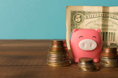 Little piggy Bank and a few stack coins with 5 dollars bill on the wooden table. Close up. The concept of saving moneyの写真素材