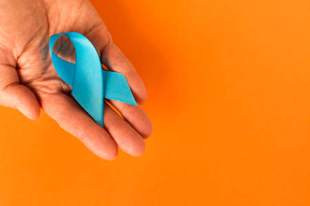 Blue ribbon in human hand on a orange background. The concept of cancer awareness. Close up. Empty...