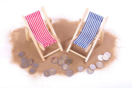 1 and 2 euro coins lie in front of toy beach chairs in the sandの写真素材