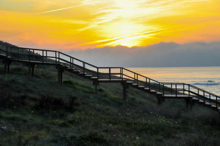 Shady silhouette of a wooden staircase from the side that leads from the ground into the sky to the sunset and in the blurry background the ocean, horizon, sky with clouds and sunの写真素材