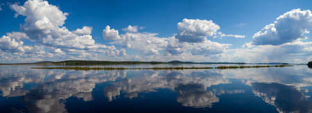 A giant panorama divided by a horizon line into the sky with large formations of clouds and a calm blue lake in which clouds are displayed. Far on the horizon you can see the evergreen forest. The waterscape is breathtaking. Plenty of air and atmosphere. Unreal nature beauty. Finland. Lapland.の写真素材