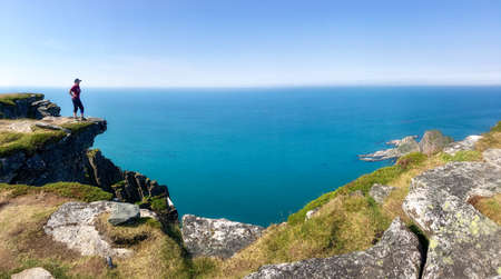 A man travels with a backpack. Hiker stands on the edge of rock and look at the background of the sea and mountains. A tourist walks along a rocky mountain trail. Norway blue sky and ocean in summer.の写真素材