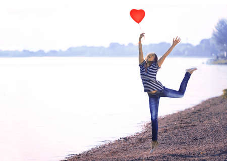 Happy Young Woman Jumping on the Shore with a Red Shaped Heart Balloonの写真素材