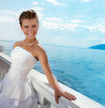 Beautiful bride wearing a fancy wedding dress, looking at camera in the sunny day at the water viewの写真素材