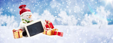 Christmas Concept: Cute Snowman Sitting with a Notice Board and a lot of Gifts on the Snow against Frost Winter Forestの写真素材