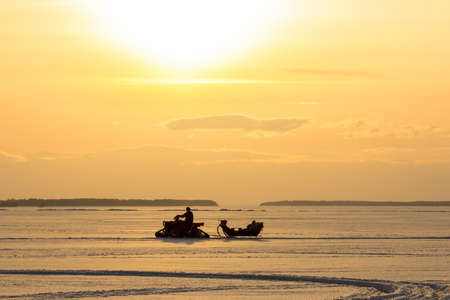 Silhouette of a man driving a snowmobile on a lake ice towing a sleigh in a sunsetの写真素材