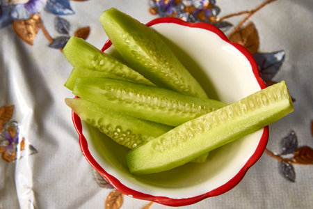 A bowl filled with freshly sliced cucumbers is sitting on a decorative table cloth, presenting a healthy and vibrant addition to the mealの写真素材