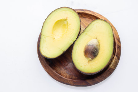 Two avocado halves are on a wooden plate with the pit removed from one. The avocado flesh is a...