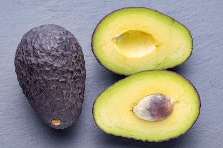 Three avocados, one whole and two halves, are arranged on a gray slate surface. The halved avocados reveal their creamy yellow flesh and large brown pits.の写真素材