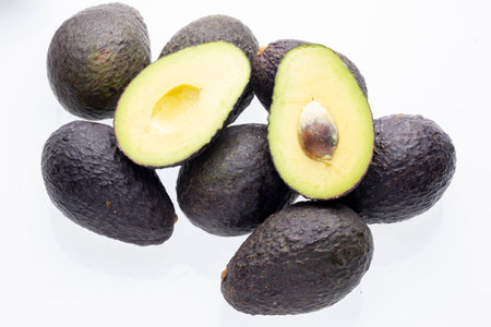 A group of six avocados, some whole and some sliced in half, with visible pits, rest on a white...