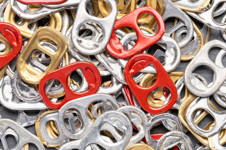 A closeup image of a variety of can pull tabs in silver, gold, and red colors scattered on a dark gray surface. The tabs are mostly silver and gold, with a few red ones interspersed. The image captureの写真素材