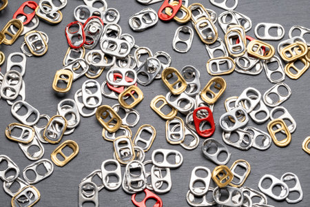 A closeup image of a variety of can pull tabs in silver, gold, and red colors scattered on a dark gray surface. The tabs are mostly silver and gold, with a few red ones interspersed. The image captureの写真素材