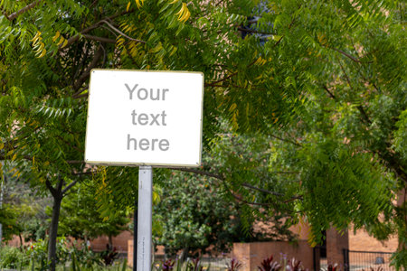 A white rectangular signboard mounted on a metal post, placed in a lush green park setting with leafy trees in the background. The sign displays a placeholder text âYour text here,â making it ideal for mockups, public message design, or customizable outdoor announcements.の写真素材