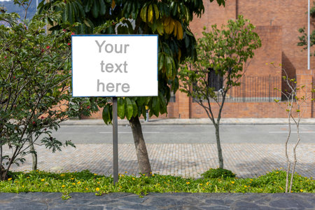 A white rectangular signboard mounted on a metal post, placed in a lush green park setting with leafy trees in the background. The sign displays a placeholder text âYour text here,âの写真素材