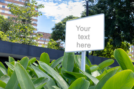 A white rectangular signboard mounted on a metal post, placed in a lush green park setting with leafy trees in the background. The sign displays a placeholder text âYour text here,âの写真素材