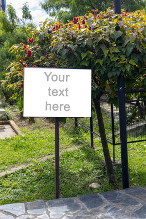 A white rectangular signboard mounted on a metal post, placed in a lush green park setting with leafy trees in the background. The sign displays a placeholder text âYour text here,âの写真素材