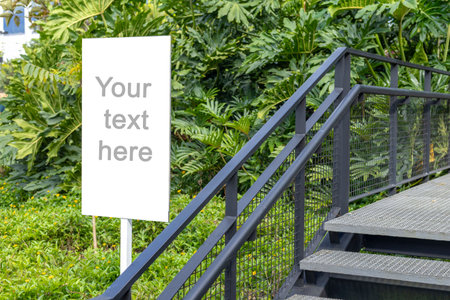A white rectangular signboard mounted on a metal post, placed in a lush green park setting with leafy trees in the background. The sign displays a placeholder text âYour text here,âの写真素材