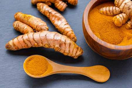 Turmeric roots and powder in a wooden bowl on a dark slate board: natural superfood and culinary spiceの写真素材