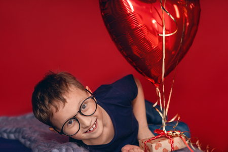 Laughting little boy in glasses, different color heart, star balloons for valentines day or birthday on red backgroundの写真素材