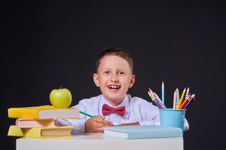 joyful little boy sitting on a black background on a table with books and educational supplies. Happy child pupil doing homework at the table.の写真素材