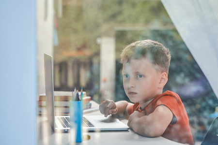 child is distracted from his studies and looks out the window. home schooling, doing homework. boy looks dreamily into the distance. little boy student sitting at a table with books and a computerrの写真素材